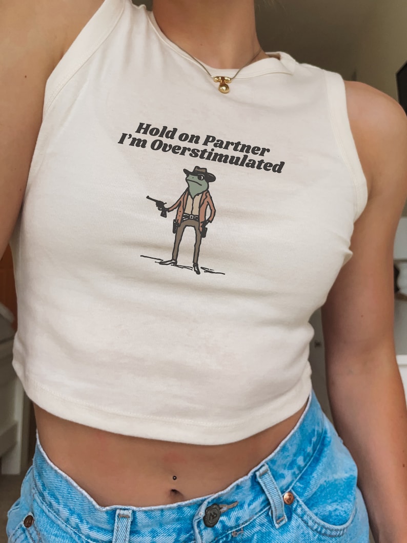 May include: Cream-colored tank top with the text "Hold on Partner I'm Overstimulated" and a cartoon frog wearing a cowboy hat and holding a gun. The frog is dressed in a brown jacket and pants. The tank top is paired with blue denim jeans.