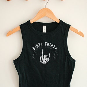 May include: Black sleeveless crop top with a white graphic print of a skeleton hand giving the middle finger with lightning bolts and the text "Dirty Thirty".
