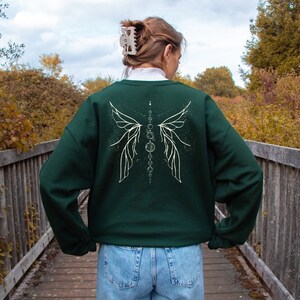 Pu&ograve; includere: Una felpa verde scuro con un disegno grafico bianco di ali e cerchi stilizzati sul retro. La felpa &egrave; abbinata a jeans blu. Lo sfondo mostra un ponte di legno e un fogliame autunnale.