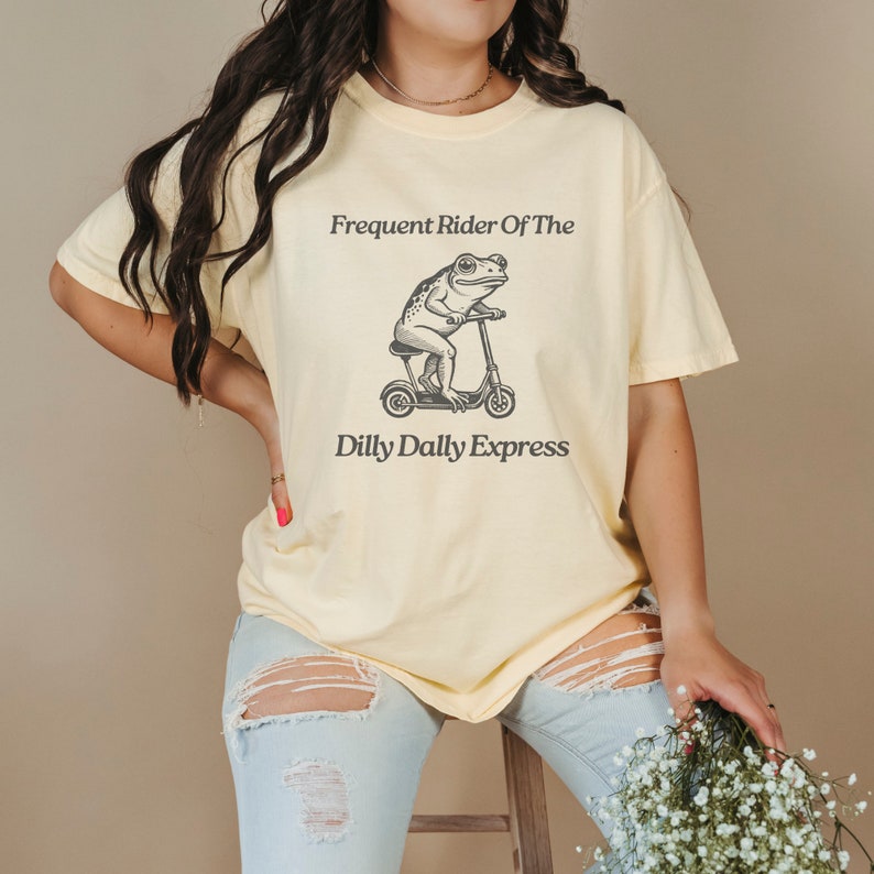 Może przedstawiać: Ż&oacute;łta koszulka z grafiką przedstawiającą żabę jadącą na hulajnodze. Tekst na koszulce brzmi "Frequent Rider Of The Dilly Dally Express".