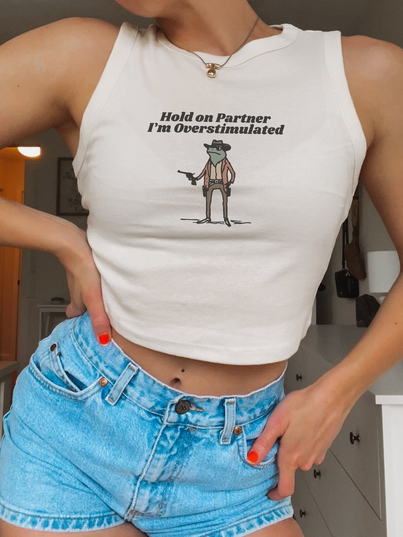 May include: Cream-colored tank top with the text "Hold on Partner I'm Overstimulated" and a cartoon frog wearing a cowboy hat and holding two guns. The tank top is paired with light blue denim shorts.