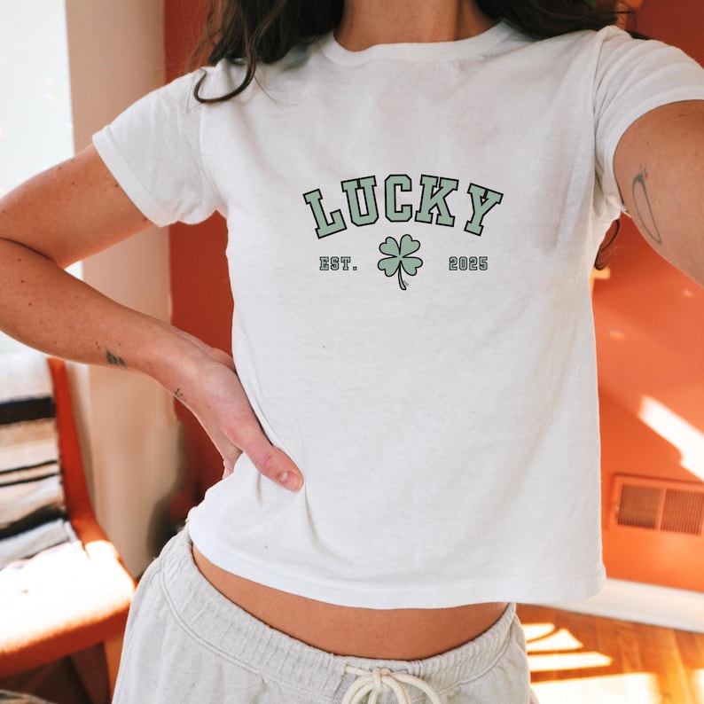 K&ouml;nnte beinhalten: Wei&szlig;es T-Shirt mit einem gr&uuml;nen vierbl&auml;ttrigen Kleeblatt-Motiv und dem Text "LUCKY EST. 2025" auf der Vorderseite.