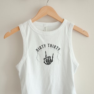May include: White sleeveless crop top with a black graphic print of a skeleton hand making a rock and roll sign with the text "Dirty Thirty" above it.