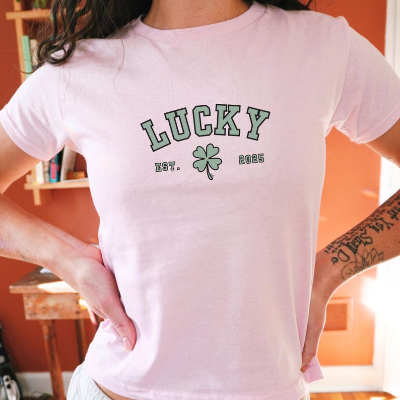K&ouml;nnte beinhalten: Ein pinkfarbenes T-Shirt mit dem Text "LUCKY" in gr&uuml;ner und schwarzer Schrift. Der Text ist von einem schwarzen Rahmen umgeben und hat darunter ein vierbl&auml;ttriges Kleeblatt. Unter dem Kleeblatt steht der Text "EST. 2025".