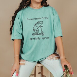 Może przedstawiać: Turkusowy t-shirt z grafiką przedstawiającą żabę jadącą na skuterze. Tekst na koszulce brzmi "Frequent Rider Of The Dilly Dally Express".