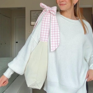 May include: A cream tote bag with a pink and white gingham bow detail. The bag is slung over the shoulder, highlighting the bow's decorative feature. The bag's simple design and the bow's pattern create a stylish accessory.