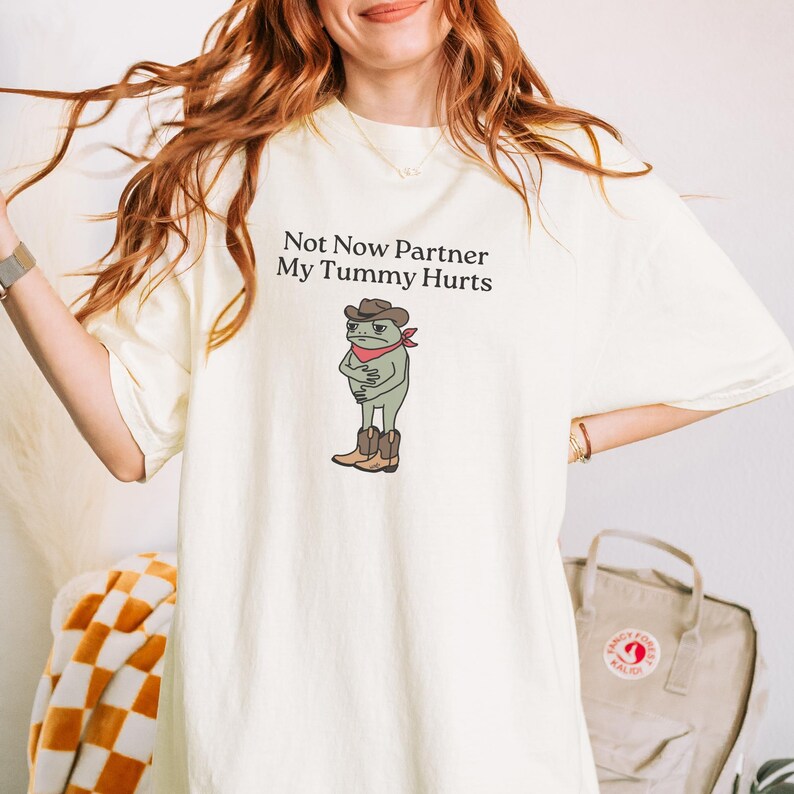 May include: Cream-colored t-shirt with the text "Not Now Partner My Tummy Hurts" and a cartoon frog wearing a cowboy hat, boots, and a red bandana. The frog is clutching its stomach. The shirt is a casual wear item.