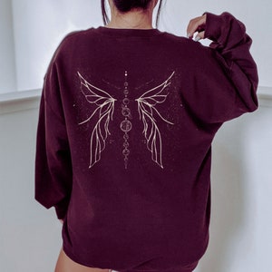 Pu&ograve; includere: Felpa bordeaux con un disegno celeste color crema sul retro. Il disegno presenta ali stilizzate e una serie di cerchi e punti interconnessi. La felpa ha maniche lunghe e girocollo.