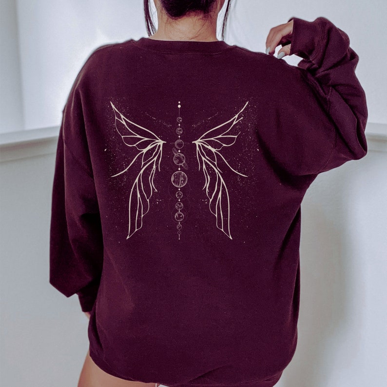 Pu&ograve; includere: Felpa bordeaux con un disegno celeste color crema sul retro. Il disegno presenta ali stilizzate e una serie di cerchi e punti interconnessi. La felpa ha maniche lunghe e girocollo.