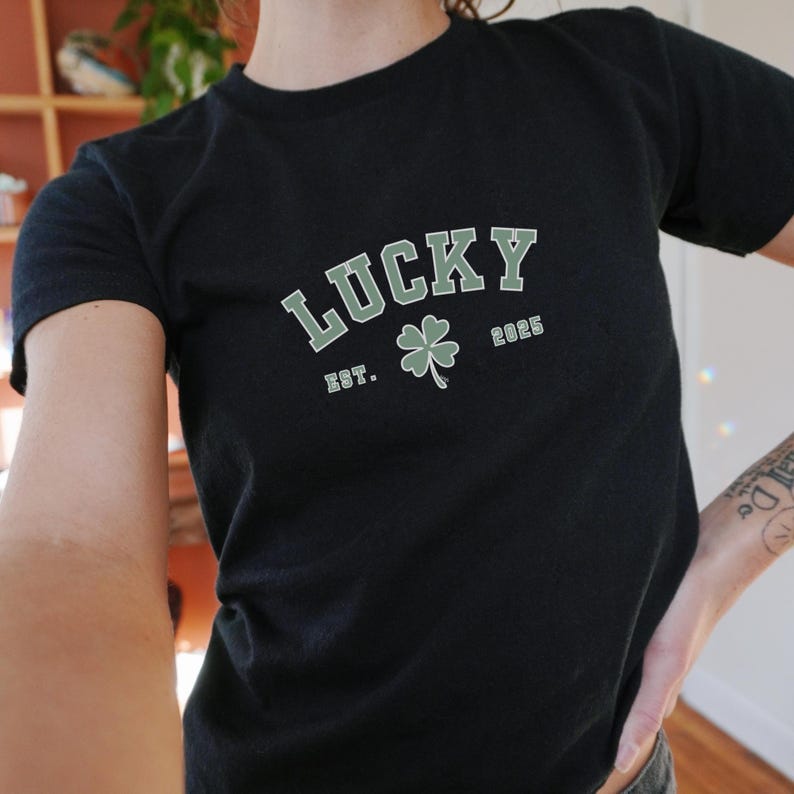 K&ouml;nnte beinhalten: Schwarzes T-Shirt mit einem gr&uuml;nen Kleeblatt und dem Text "LUCKY EST. 2025" in Wei&szlig;.