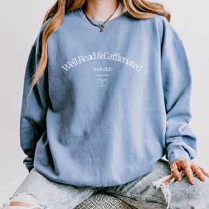 Puede incluir: Una sudadera azul claro con el texto "Well Read & Caffeinated book club Established 2019" en blanco.