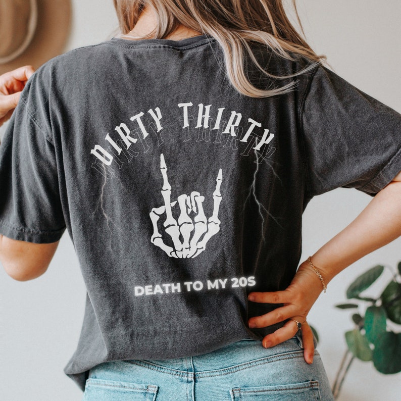 May include: Dark gray t-shirt with the text "DIRTY THIRTY" in a gothic font above a white skeleton hand making a rock and roll gesture. Below the hand is the text "DEATH TO MY 20S".