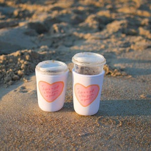 May include: Two white coffee cup sleeves with pink heart designs and the text "Life's a gift & the giver is good!" are on a sandy beach. The cups have clear lids and are filled with a dark liquid.