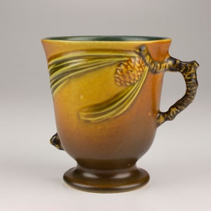 May include: A brown and green ceramic mug with a pine cone and pine needles design. The mug has a handle shaped like a pine branch.
