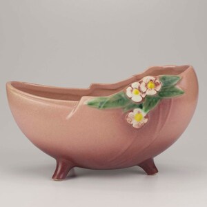 May include: A vintage, pink ceramic planter with three small legs. The planter has a curved, boat-like shape and is decorated with a green leaf and white flower design. The planter is likely from the mid-20th century.