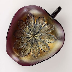 May include: A dark purple glass bowl with a handle. The bowl has a gold iridescent finish and a detailed floral design.