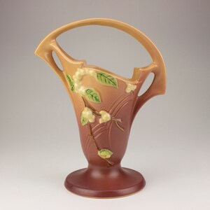 May include: A brown and tan ceramic vase with a handle and a floral design. The vase has a wide base and a narrow neck.