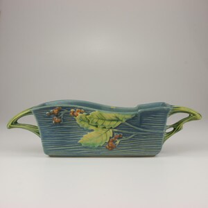 May include: A rectangular ceramic planter with a blue-toned, wood-grain textured surface. The vessel features relief-molded green holly leaves and brown berries on the front, with two stylized, branch-like handles extending from the sides.