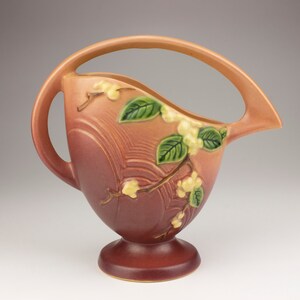 May include: A vintage ceramic pitcher with a unique, curved design. It features a pink glaze with a floral pattern of white blossoms and green leaves.