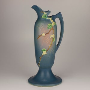 May include: A tall, blue ceramic pitcher with a handle and a flared base. The pitcher is decorated with a branch design featuring white flowers and green leaves. The pitcher has a unique spout design.