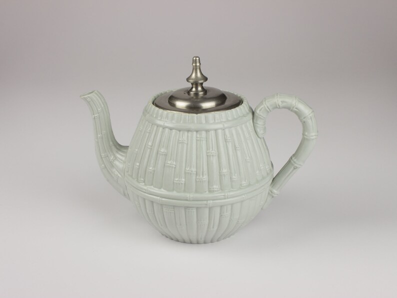 May include: A light green ceramic teapot with a silver lid and a bamboo-like design. The teapot has a round body and a curved spout.