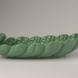 May include: A green ceramic serving dish shaped like a leaf with a curled handle. The dish is glazed and has a smooth, glossy finish.