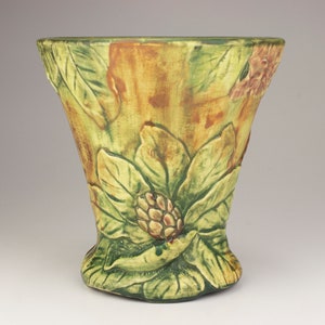 May include: A green and brown ceramic vase with a floral design. The vase has a wide mouth and a narrow base. The floral design features large leaves and a flower with a brown center.