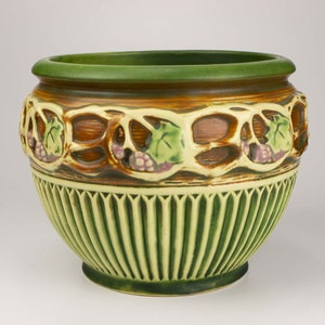 May include: A ceramic planter with a green rim and base, featuring a decorative band of grapes and leaves. The planter has a light green, ribbed lower section and a brown and tan upper section with grape and leaf cutouts.