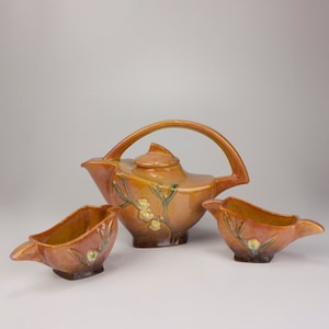 May include: A brown ceramic teapot and two matching creamer or sugar bowls with a floral design. The teapot has a unique curved handle and a lid with a knob. The bowls have a rounded shape with a spout and a handle.