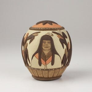 May include: A ceramic sphere with a brown, white, and orange design. The sphere features a relief image of a woman with long dark hair and a corn stalk design around the sphere.