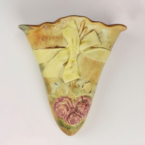 May include: A ceramic wall vase with a yellow bow and pink roses. The vase is a light brown color with a textured surface.