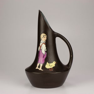 May include: A dark brown ceramic pitcher with a unique shape and a painted design of a boy and a chick. The pitcher has a handle and a wide base.