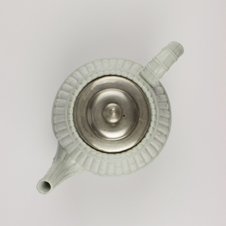 May include: A light blue ceramic teapot with a silver lid and a decorative design around the top. The teapot has a spout and a handle.