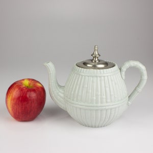 May include: A light blue teapot with a silver lid and a bamboo design. The teapot is sitting on a white surface next to a red apple.