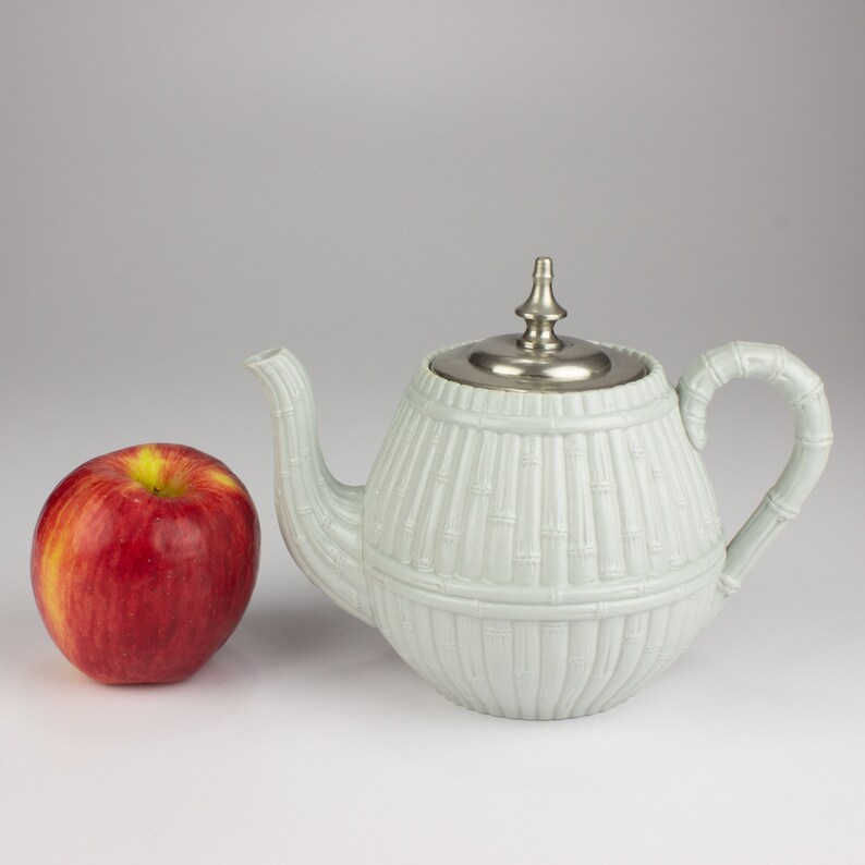 May include: A light blue teapot with a silver lid and a bamboo design. The teapot is sitting on a white surface next to a red apple.