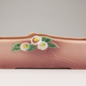 May include: A pink ceramic planter with a floral design. The planter has a rectangular shape with rounded edges and features white flowers and green leaves.