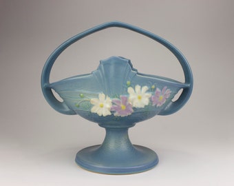Puede incluir: Una cesta de cerámica azul con asa y diseño floral. La cesta está sobre una base de pedestal.