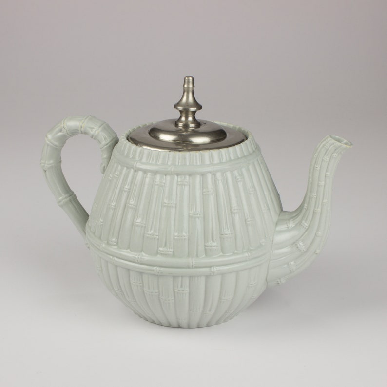 May include: A light green ceramic teapot with a silver lid and a bamboo design. The teapot has a round body and a curved spout.