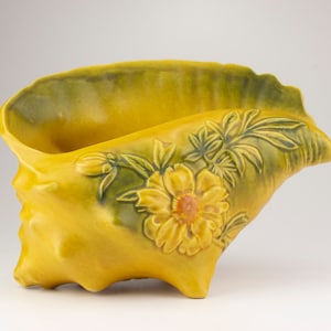 May include: A yellow ceramic seashell planter with green and yellow floral details. The planter is shaped like a conch shell and has a wide opening at the top.