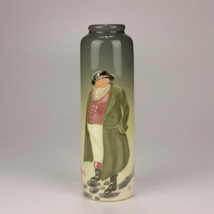 May include: A tall, cylindrical ceramic vase with a green glaze. The vase features a painted image of a man in a green coat, red vest, and white pants standing on a path with stones.