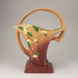 May include: A brown and tan ceramic vase with a ring handle and a floral design. The vase has a white and green floral design with a brown and tan background.