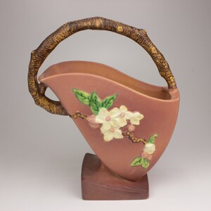 May include: A pink ceramic vase with a brown twig handle and a floral design of white blossoms and green leaves.