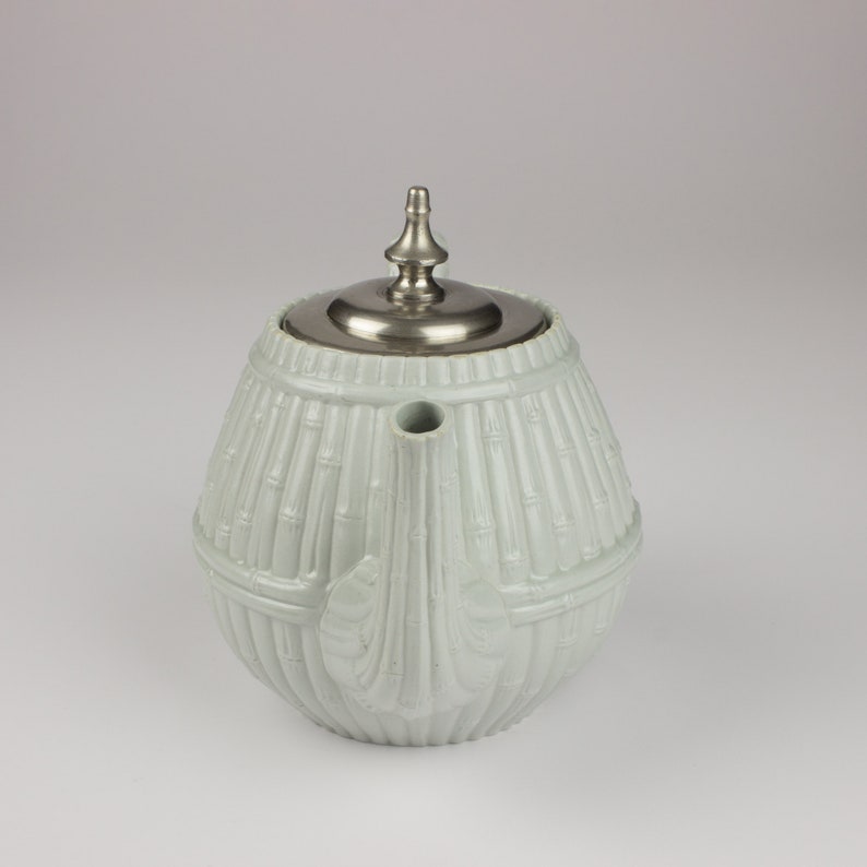 May include: A white ceramic teapot with a silver lid and a bamboo-like design. The teapot has a rounded body and a spout on the side.