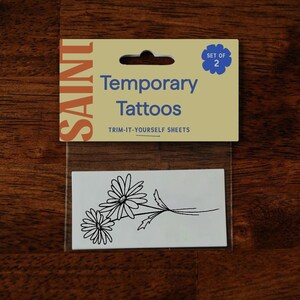Pu&ograve; includere: Un pacchetto di tatuaggi temporanei con un disegno a margherita. La confezione recita "SAINT Temporary Tattoos" e "TRIM-IT-YOURSELF SHEETS". Il set include due fogli di tatuaggi.