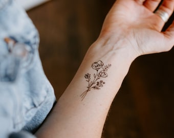 Puede incluir: Un tatuaje temporal de un delicado ramo floral, con amapolas y otras flores silvestres, se muestra en un brazo. El fino arte lineal está en tinta marrón, creando una estética natural y elegante. El brazo está frente a un fondo borroso.