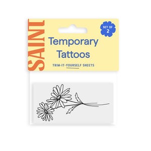 Pu&ograve; includere: Un pacchetto di tatuaggi temporanei con un disegno di margherite in bianco e nero. La confezione include una scheda gialla e bianca con le parole "Temporary Tattoos" e "Trim-It-Yourself Sheets". La scheda ha anche la parola "SAINT" in arancione.