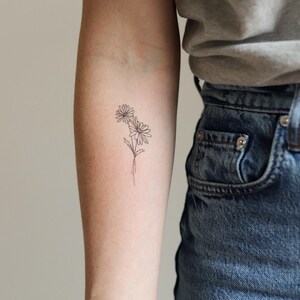 Pu&ograve; includere: Un tatuaggio minimalista a contorno nero di due margherite su un braccio. Le margherite hanno petali dettagliati e sono collegate da sottili steli. Il tatuaggio &egrave; sull'avambraccio, con una maglietta grigia e jeans blu sullo sfondo.