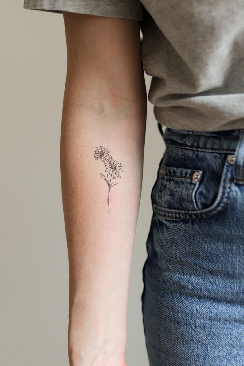 Pu&ograve; includere: Un tatuaggio minimalista a contorno nero di due margherite su un braccio. Le margherite hanno petali dettagliati e sono collegate da sottili steli. Il tatuaggio &egrave; sull'avambraccio, con una maglietta grigia e jeans blu sullo sfondo.