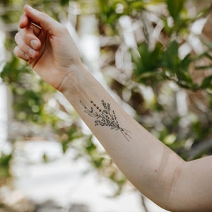 May include: A black and white floral tattoo design on the forearm. The tattoo features a delicate bouquet of leaves and small flowers tied together with a simple line. The background is blurred with green foliage.