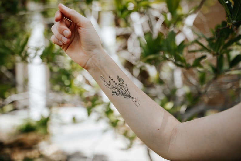 May include: A black and white floral tattoo design on the forearm. The tattoo features a delicate bouquet of leaves and small flowers tied together with a simple line. The background is blurred with green foliage.